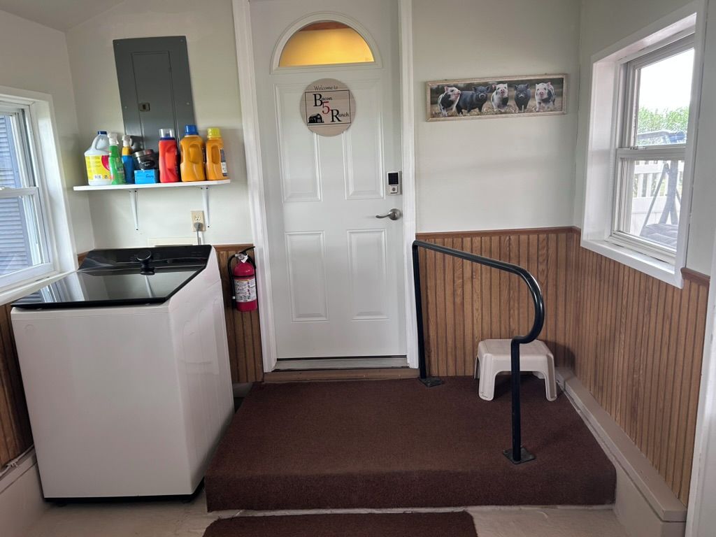 A kitchen with a refrigerator, a white door with a welcome sign, a shelf with cleaning supplies, and a window with a view.