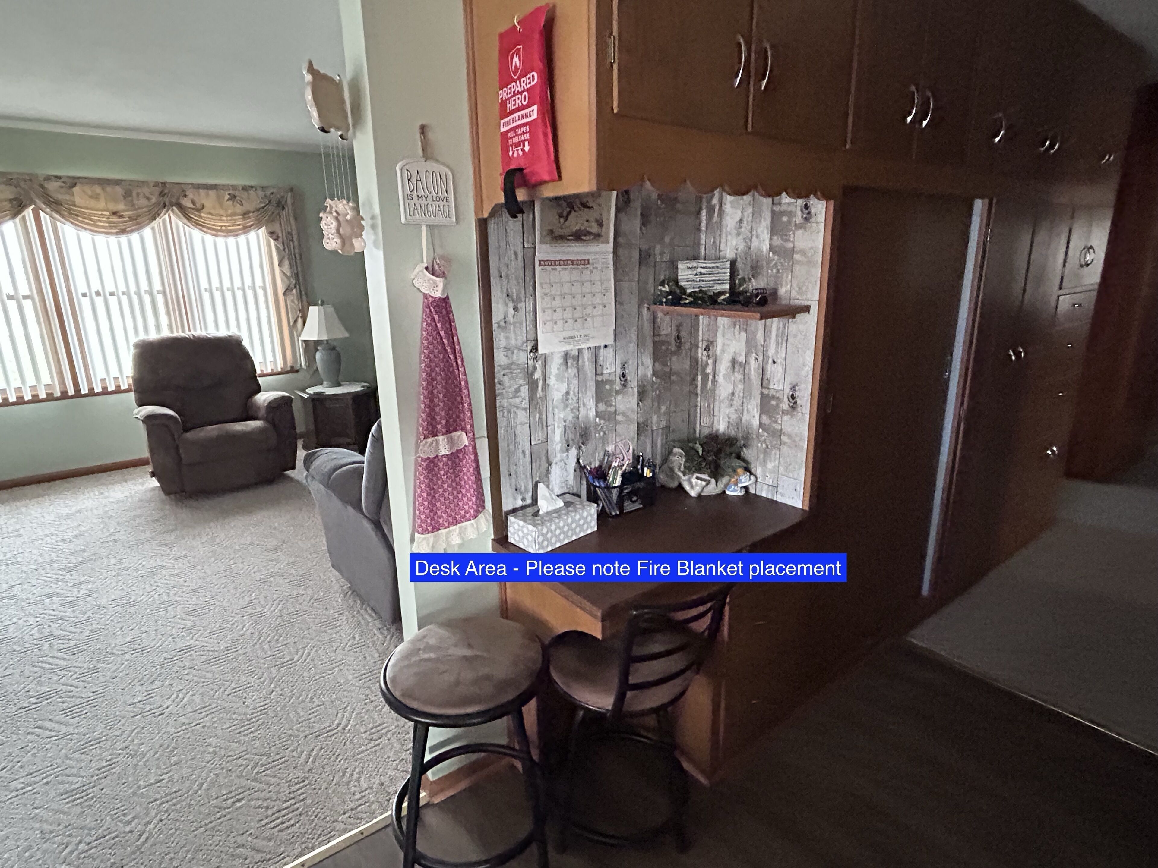 A kitchen area with a fire blanket hanging on the wall, a chair, a table with stools, and a window with blinds.