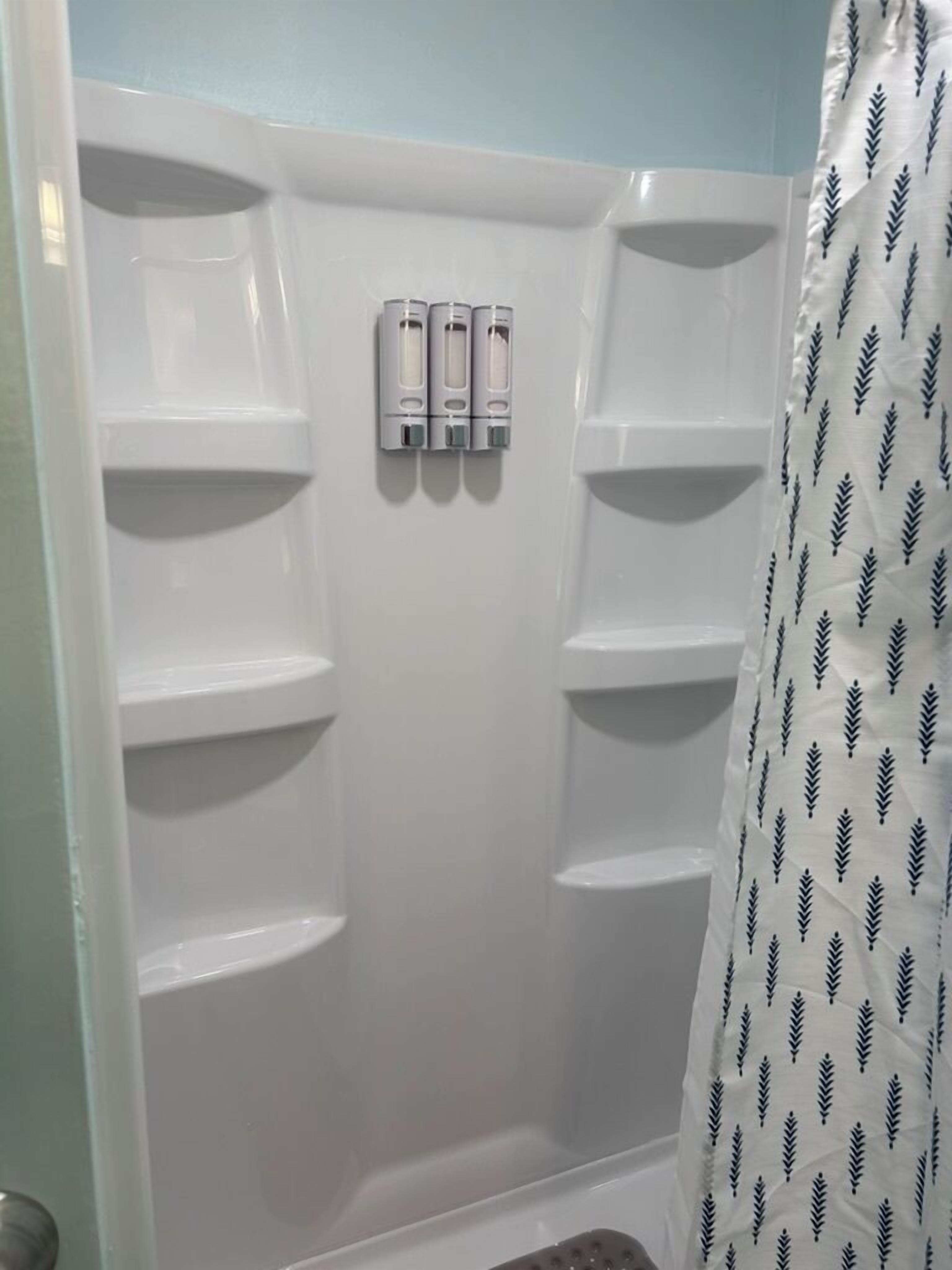 Zona de ducha con tres botellas de champú y acondicionador en la pared, cortina de baño con diseño de hojas y suelo de baldosas.