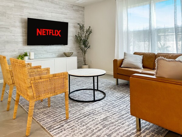 A modern living room with a flat-screen TV mounted on a wall, a brown leather sofa, a round coffee table, wicker chairs, and a potted plant.