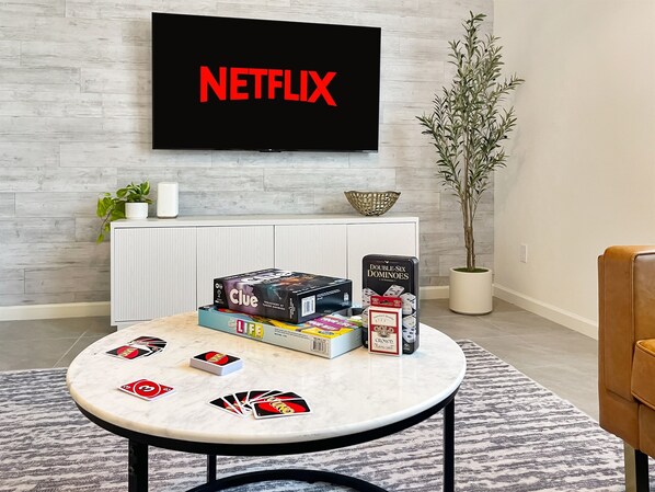 Sala de estar com televisão exibindo 'NETFLIX', um armário branco, uma planta e uma mesa com jogos de tabuleiro.