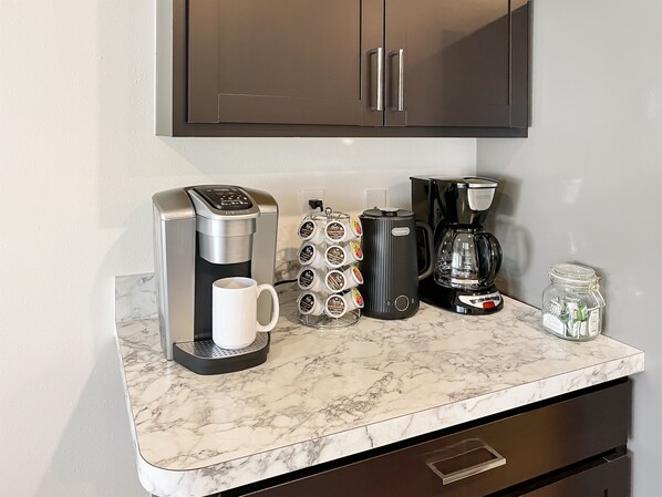 Una encimera de cocina con cafetera, jarra de café, taza, tarro de té y molinillo de café.