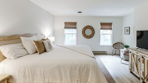 Un dormitorio con una cama grande, dos ventanas con persianas, una televisión y una silla de mimbre.