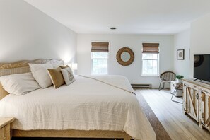 Ein Schlafzimmer mit einem großen Bett, zwei Fenstern mit Jalousien, einem Fernseher und einem Korbstuhl.