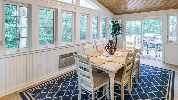 Un comedor con techo de madera, ventanas amplias, una mesa con sillas y un tapete azul estampado.