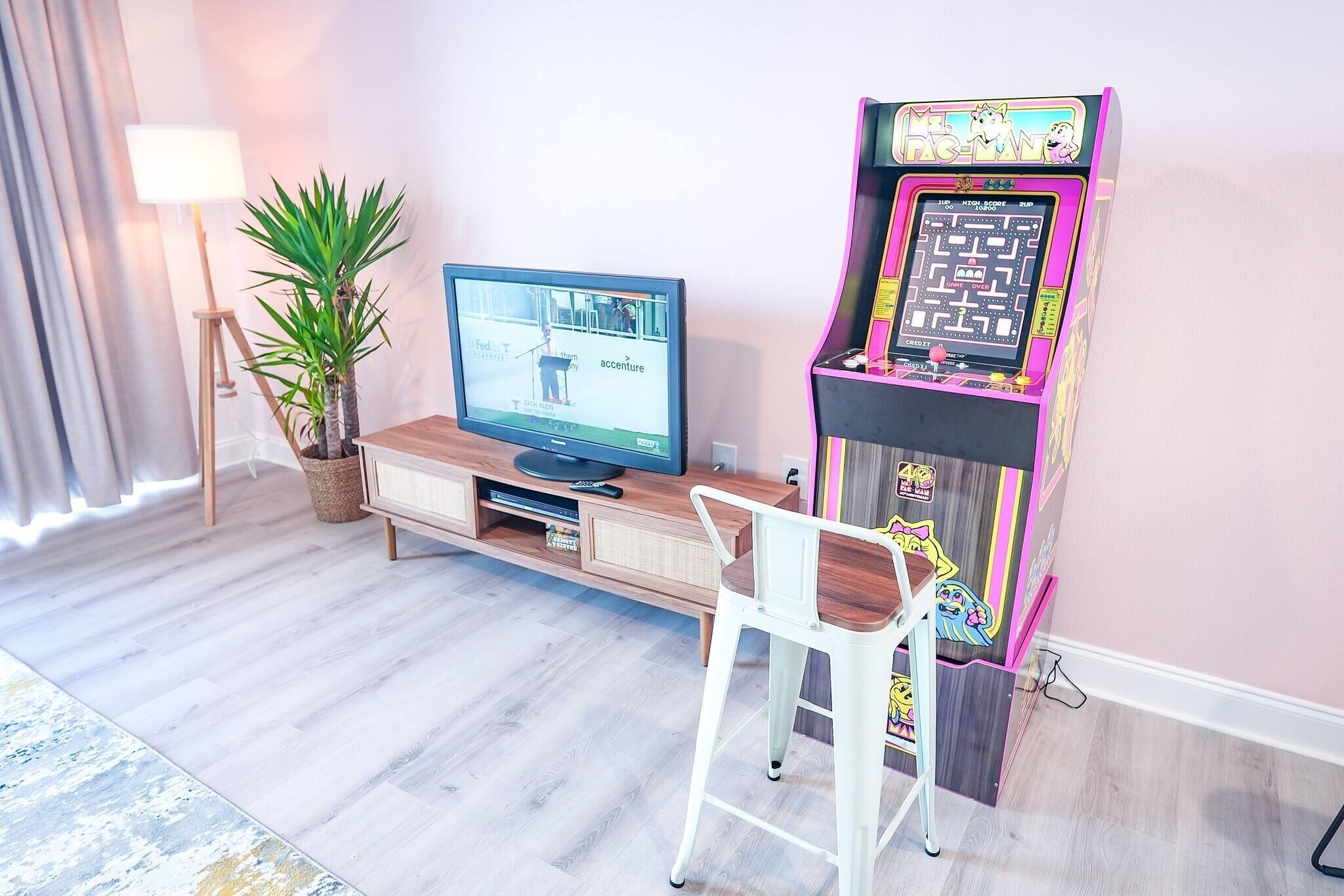 Une pièce comprenant un téléviseur, une borne d’arcade, une plante en pot, une lampe et une chaise blanche.