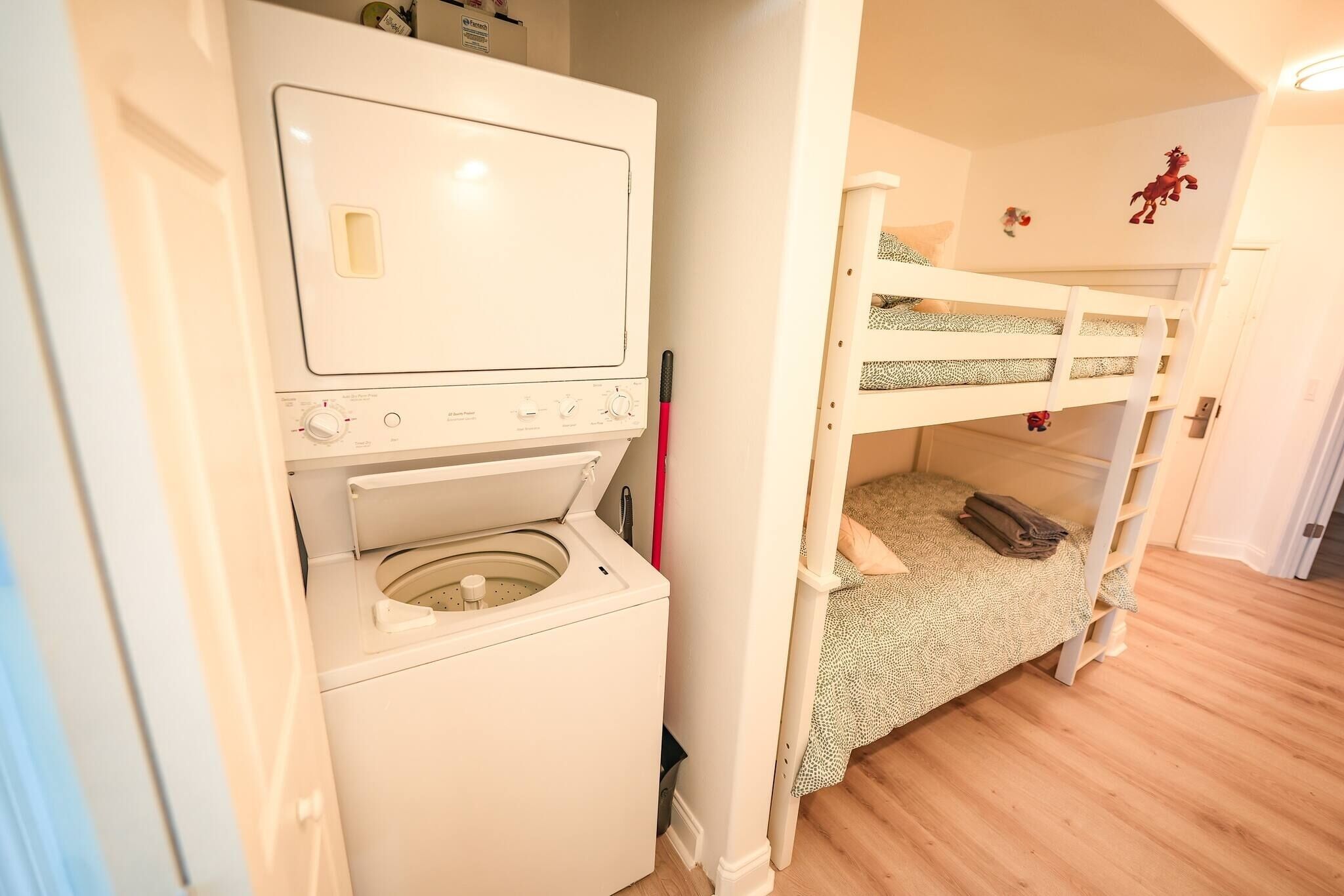 Un área de lavandería con lavadora y secadora, una cama litera con cubrecama estampado en verde, y un juguete rojo sobre la cama.