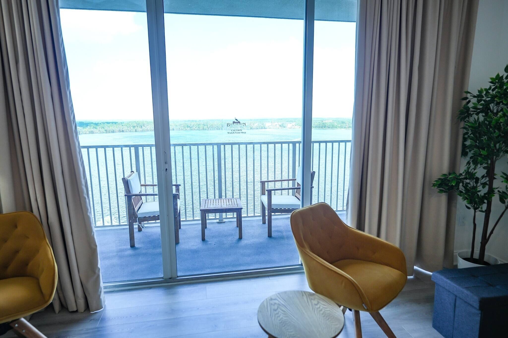 A room with a view of the sea, a balcony with chairs, and a plant.