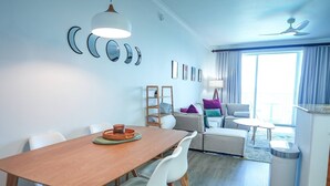 Un coin repas avec une table en bois et des chaises blanches, un canapé avec des coussins, une petite table avec une lampe et une poubelle.