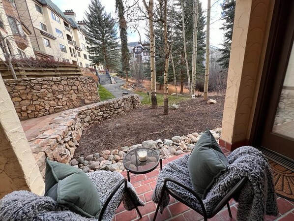 Eine Terrasse mit zwei Stühlen, einem Tisch, einer Steinmauer und Blick auf ein Gebäude und Bäume.