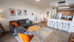 A modern living room with a grey sectional sofa, orange armchairs, a wooden coffee table, and a dining area with a white table and chairs.