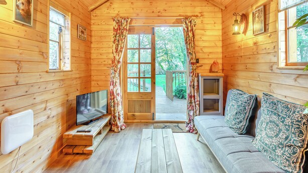 L'intérieur d'un chalet en bois chaleureux et accueillant, avec un téléviseur posé sur un meuble en bois, un canapé agrémenté de coussins à motifs et une porte en bois donnant sur une terrasse extérieure.