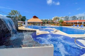 A swimming pool with a waterfall feature, lounge chairs, and a covered area with a red roof. - Villas Paraíso Coco 55 (Jacó)