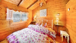 Un dormitorio de cabaña de madera con una cama, dos mesitas de noche, una ventana con cortinas de encaje, un reloj de pared y un cuadro enmarcado.