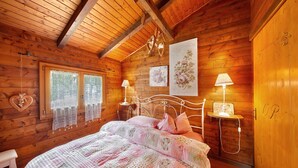 A wooden cabin bedroom with a bed, two bedside lamps, a window with curtains, and a wall with a floral painting.