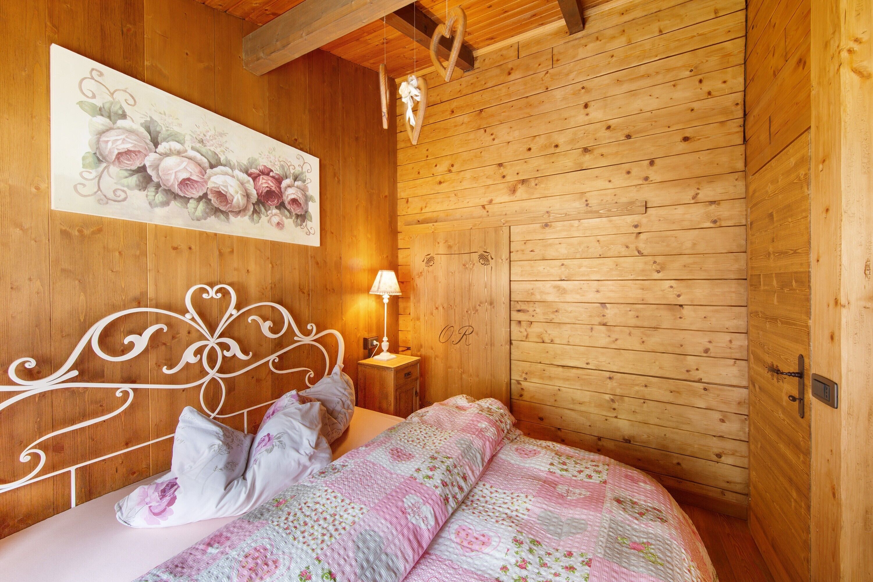 Een kamer met houten lambrisering, een bed, een nachtkastje, een wandlamp en een bloemrijk schilderij.
