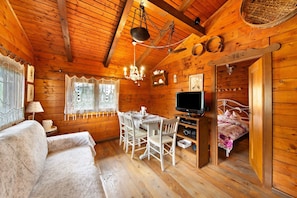 A wooden cabin interior with a dining table, chairs, a television, a bed, and a chandelier.