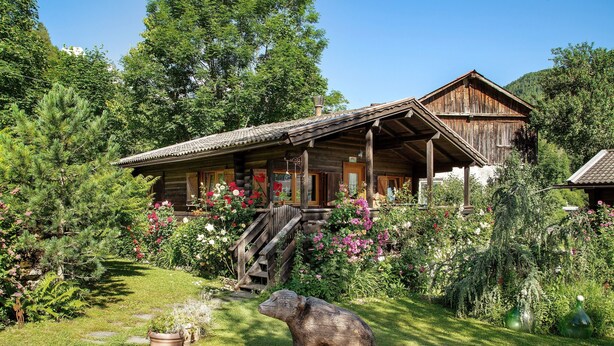 Een houten chalet met veranda, omringd door weelderige begroeiing en bloeiende planten.