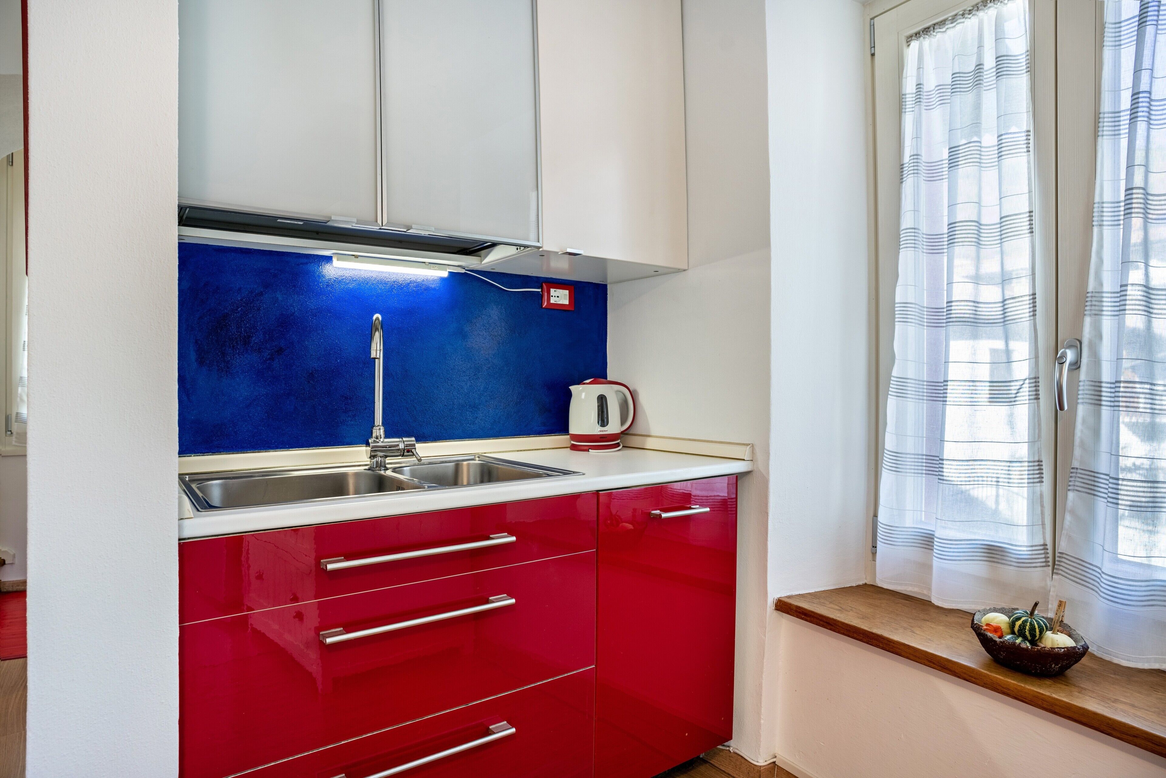 Une cuisine avec des meubles de couleur rouge, un évier en acier inoxydable et une bouilloire posée sur le plan de travail.