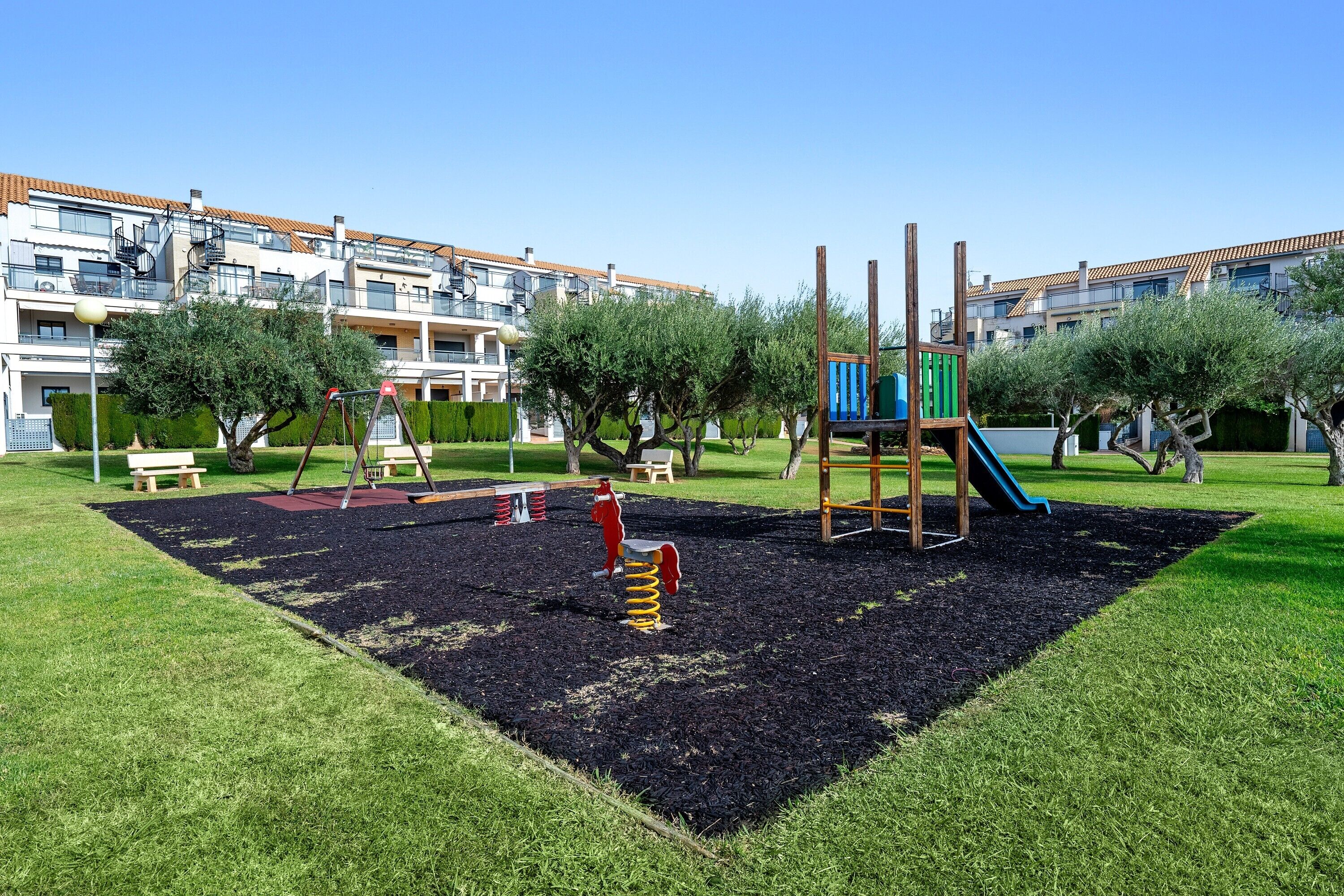 Un terrain de jeux avec un toboggan et des balançoires, entouré de pelouse et d’arbres, avec des immeubles d’habitation en arrière-plan.