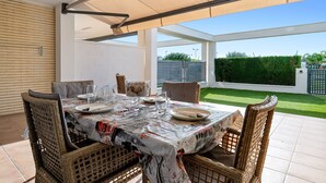 Un espace extérieur abrité, aménagé pour les repas, avec des meubles en osier, une table dressée pour deux personnes et une vue sur un jardin bien entretenu.