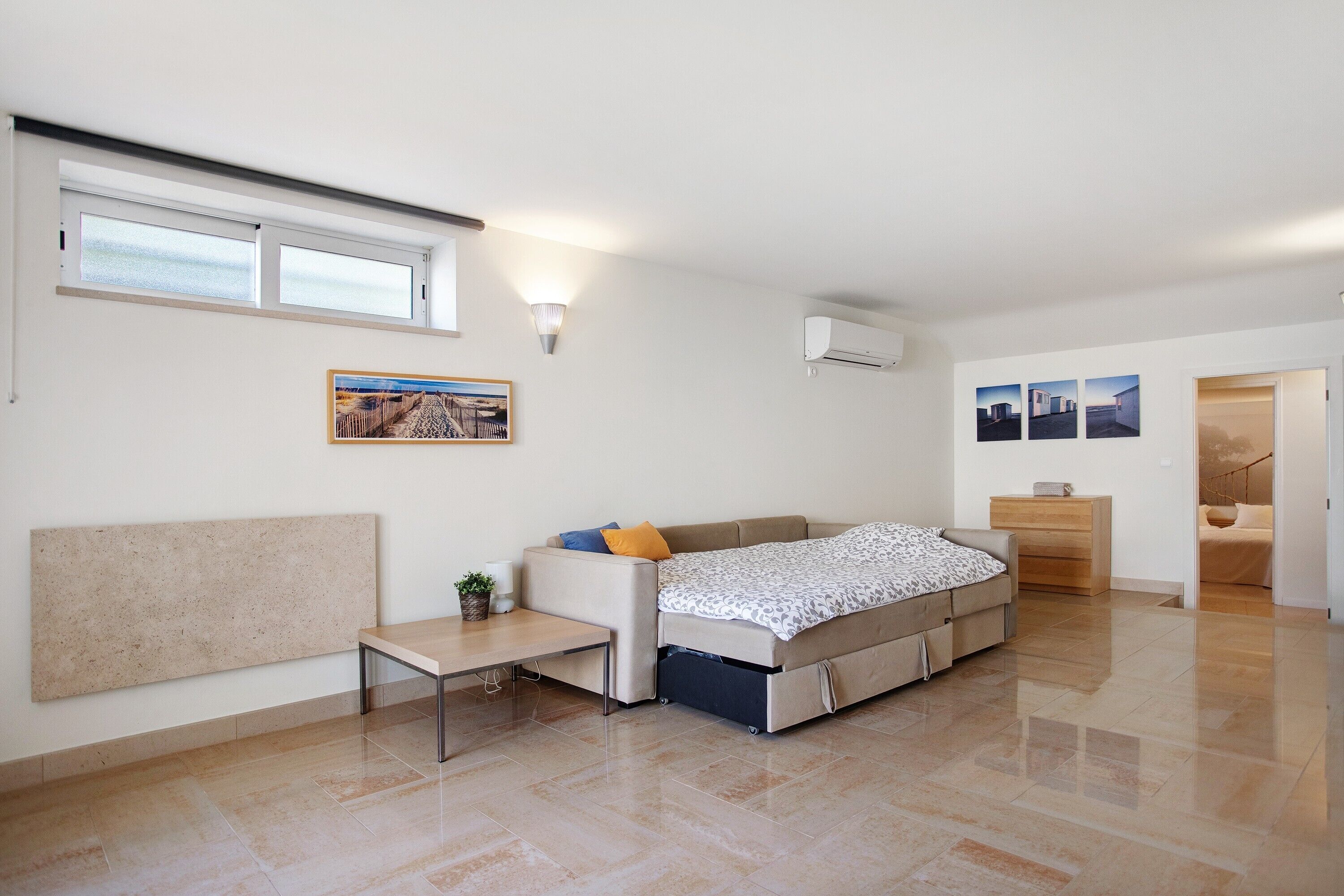 Une chambre moderne et bien éclairée, équipée d’un lit, d’une petite table, d’un climatiseur fixé au mur et d’une fenêtre.