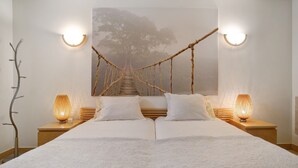 En säng med ett vitt överkast, två sänglampor, en sänggavel i trä och en väggdekoration i form av ett repbro.