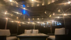 An outdoor seating area with a central table and cushioned chairs, surrounded by a dark, curved ceiling with string lights.