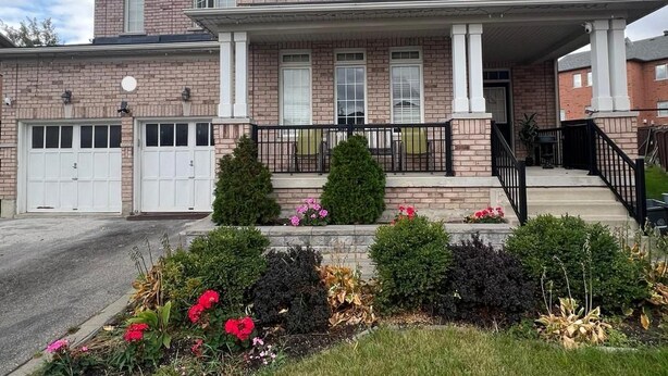 A two-story brick house with a front porch, white garage doors, and a landscaped front yard with various shrubs and flowers.