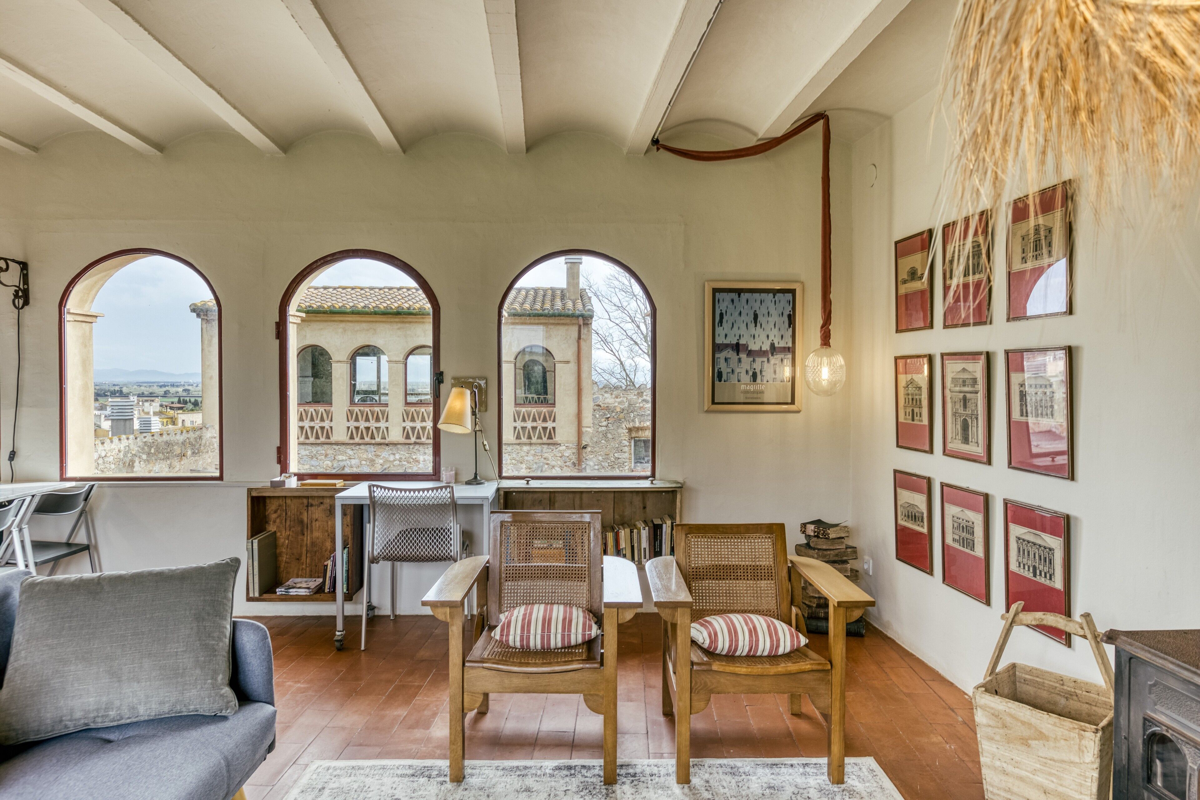 A room with a sofa, two chairs, a table, and a fireplace. There are three arched windows with views of buildings and a cityscape.