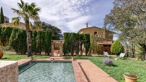 Una piscina con borde de azulejos, rodeada por un camino de ladrillo, y un edificio con techo de tejas al fondo.