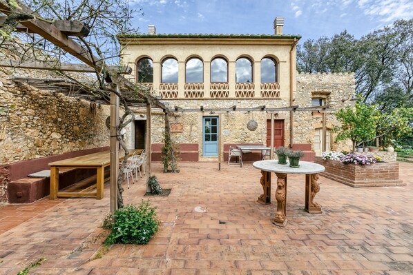 A stone building with arched windows, a wooden pergola, and outdoor seating.