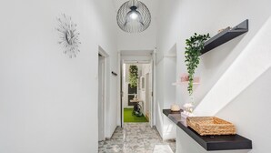 Un pasillo con suelo de baldosas, un lámpara colgante, un estante con una planta y una puerta que da a otra habitación.
