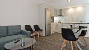 Una cuina moderna amb nevera, forn i fogó, una zona de menjador amb taula de vidre i cadires, i un espai de sala d'estar amb sofà i cadires.