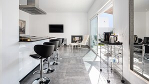 A modern kitchen with a flat-screen TV, bar stools, a coffee machine, and a glass-enclosed dining area.