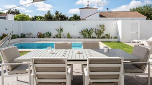 A poolside patio with a table and chairs, surrounded by a white wall and greenery.