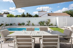 A poolside patio with a table and chairs, surrounded by a white wall and greenery.