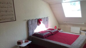 A bedroom with a bed, a nightstand, a red lamp, a window, and a wall with a framed picture. - Apartment 'Le Grenier D'annick' with Mountain View, Private Terrace and Wi-Fi (Villefort)