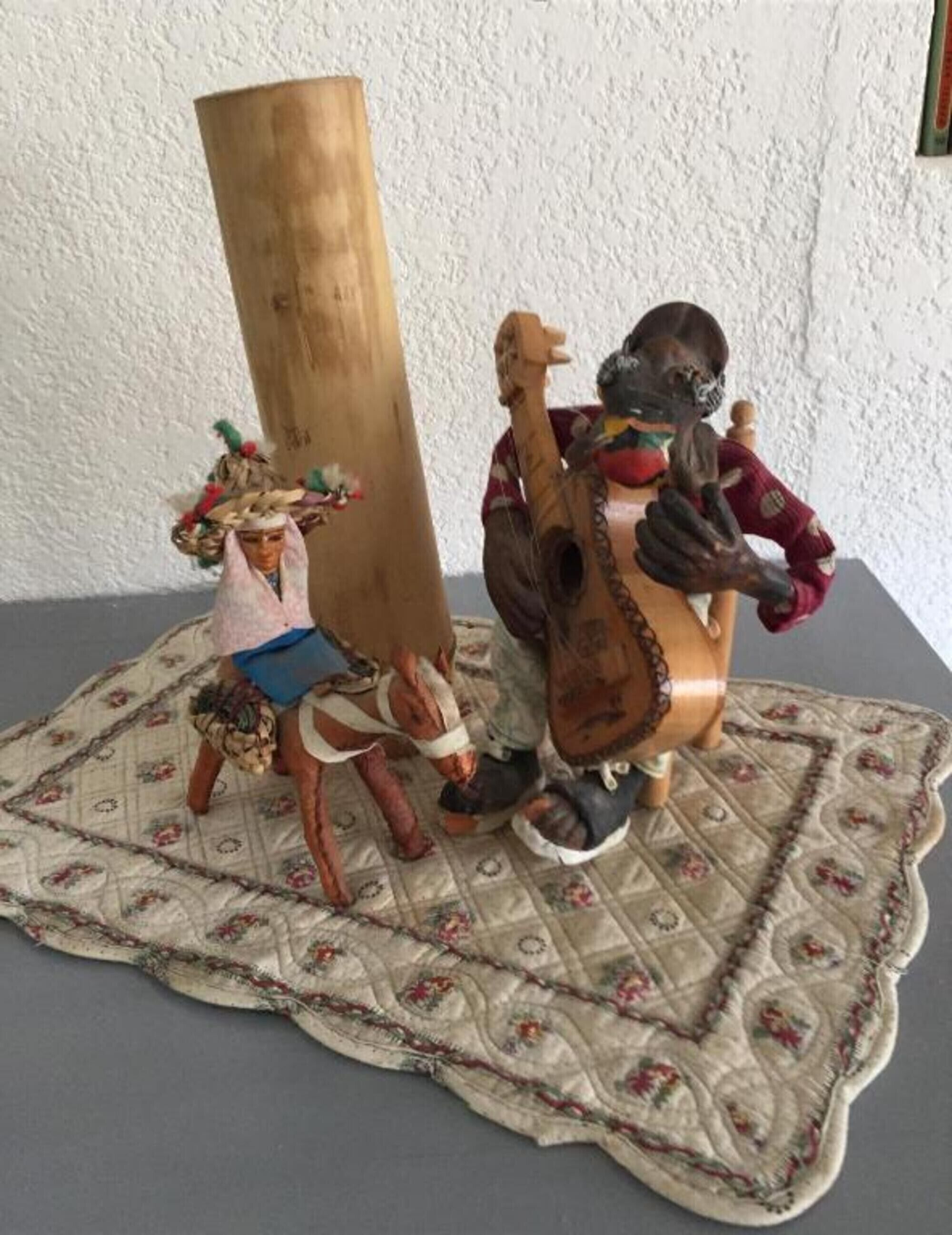 A small figurine of a person playing a guitar, a tall wooden object, and another figurine with a basket on their head.
