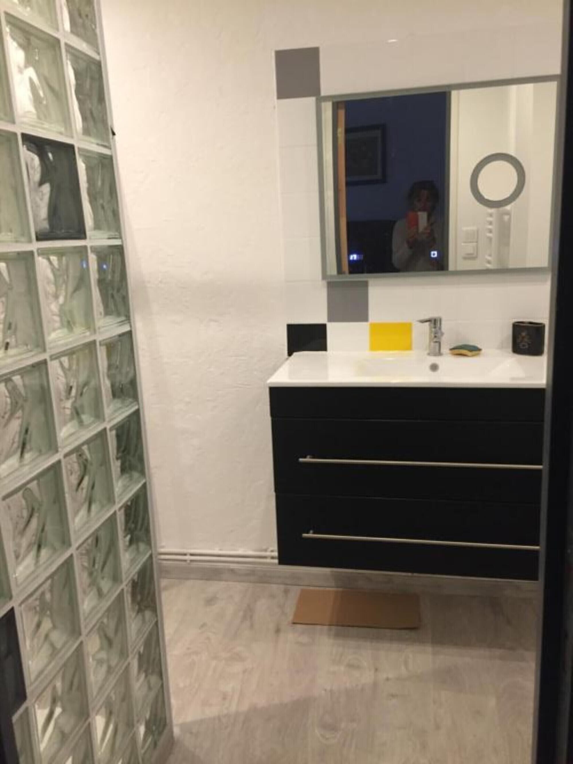Une salle de bain avec une douche à parois de verre, un lavabo blanc avec un distributeur de savon jaune et un meuble-lavabo noir doté de tiroirs.