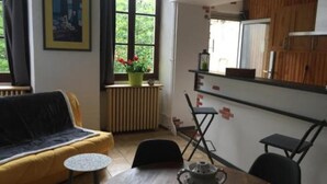 Een knus woongedeelte met een bank, een klein rond tafeltje, een radiator en een raam met uitzicht op groen.