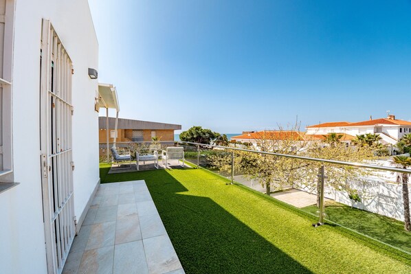 Un balcón residencial con vistas a un césped verde, un edificio blanco y un cielo despejado.