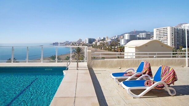 A poolside area with lounge chairs, a clear view of the sea, and a cityscape in the background.