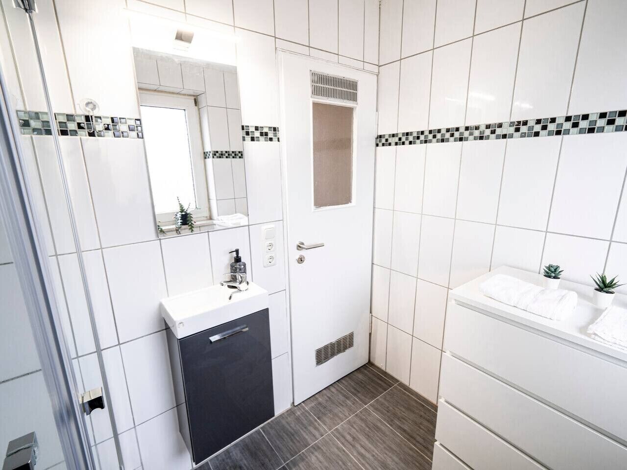 Ein modernes Badezimmer mit weißen Fliesen, einem Spiegel, einem Waschbecken und einer Duschkabine.