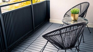 Ein Balkon mit zwei schwarzen Drahtstühlen, einem kleinen runden Glastisch und einem goldenen Topf mit Grünpflanzen.