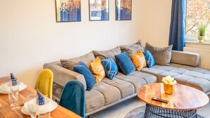 Ein modernes Wohnzimmer mit grauem Sofa, bunten Kissen, einem Holztisch und drei gerahmten Kunstwerken an der Wand.