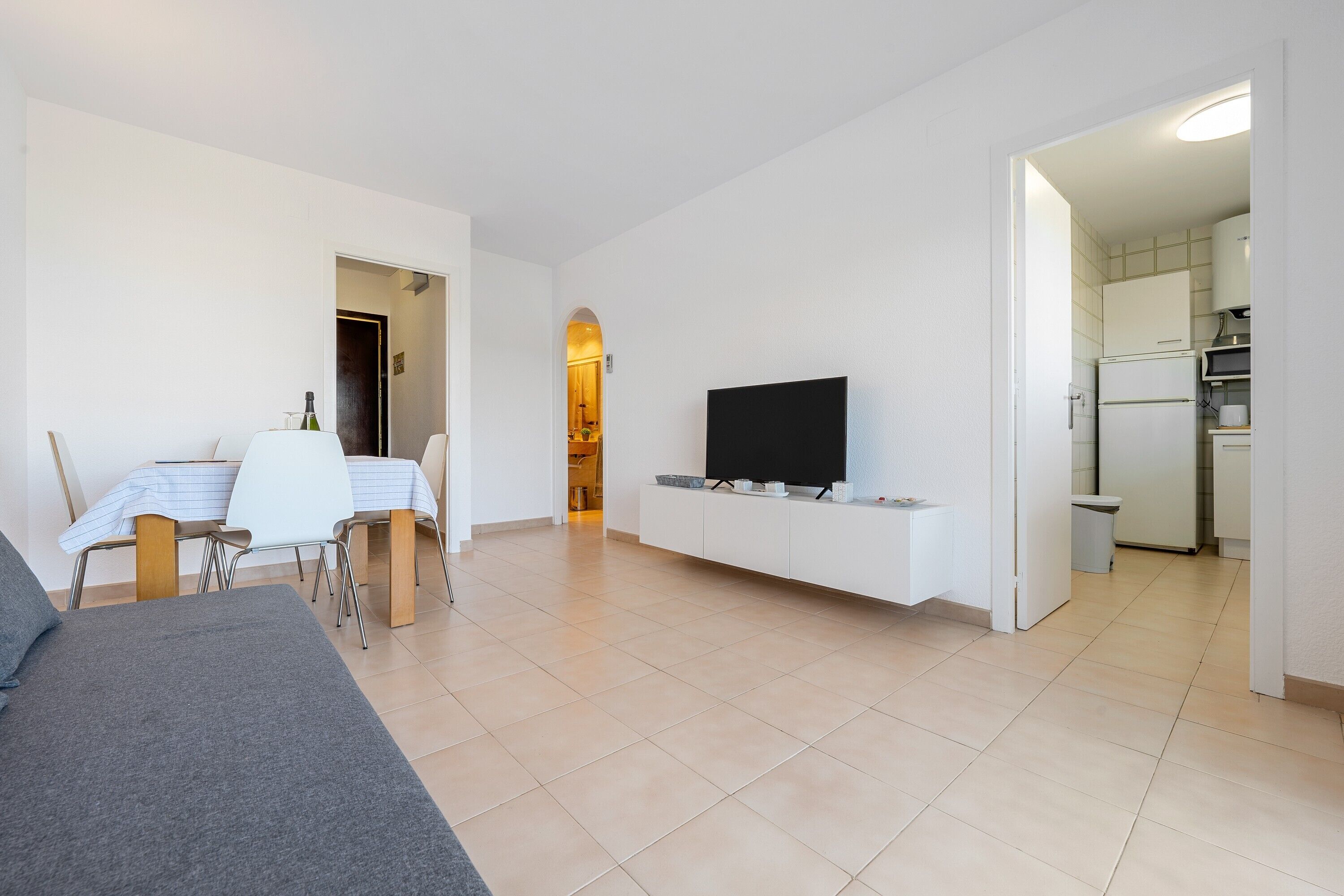 Un salon moderne comprenant un canapé, une table à manger, des chaises, un téléviseur à écran plat et un coin cuisine visible à travers une porte ouverte.