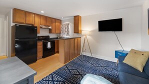 A compact kitchen with wooden cabinets, a black refrigerator, and a stove. A blue patterned rug covers the floor, and a blue sofa is in the living area. - Good 2 Go at L Rancho Court (Spearfish)