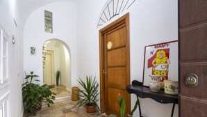 Un pasillo con una puerta de madera, plantas en macetas, un estante negro con utensilios de cocina y un cartel en la pared.