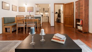 Een woonkamer met een houten eettafel, stoelen, een boekenkast en een deur naar een keuken.